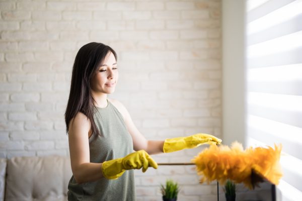 young woman dusting television top wearing yellow gloves with orange duster
